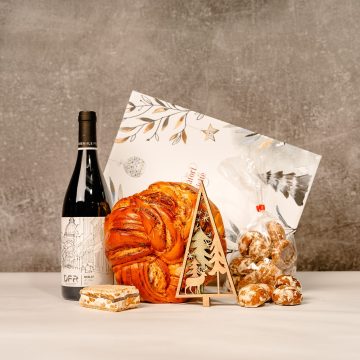 in fundal se vede o cutie alba de cadou, in prim plan e o sticla de vin rosu, cozonac rotund, turta dulce cu glazura de vanilie, halvita cu ciocolata si o decoratiune - brad de lemn cu 3 straturi. se vede o vulpe si doi brazi unul culoarea lemnului, unul verde