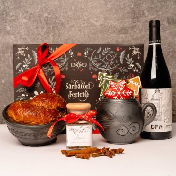 in prim plan se vede un castron negru in care este un cozonac, un borcan de sticla cu funda rosie si condimente de vin fiert, cana din ceramica neagra in care sunt 3 bucati de turta dulce, model brad verde, steluta rosie si brad natur, stila de vin rosu cu eticheta alba, crama DFr, in fundal se vede o cutie pentru cadou cu grafica neagra si arabescuri albe cu dunda rosie, asortata cu funda de la borcanul de condimente